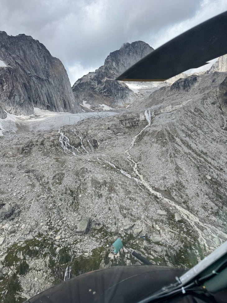 An aerial view of the green-roofed Kain Hut, Snowpatch Spire (left), and Bugaboo Spire. The rightmost "waterfall" does not typically exist, and signals the high water level in the Kain Hut Basin at the moment.
