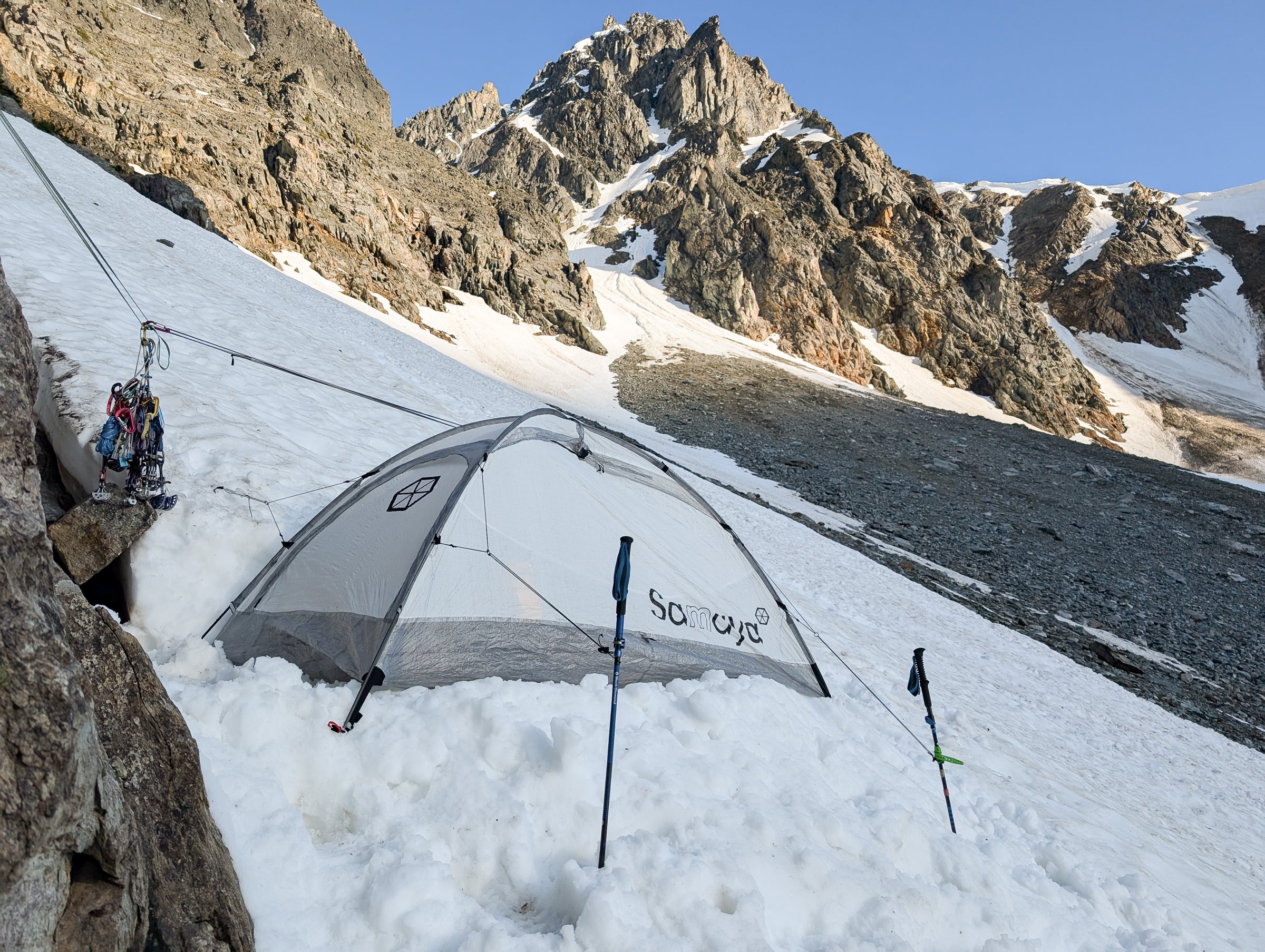 A Samaya Assaut2 climbing tent is pitched on a steep snow slope in a mountain valley.